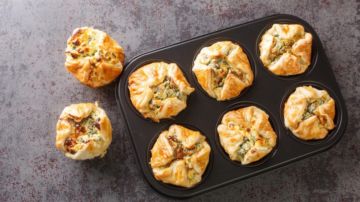 Blätterteig-Muffins mit Rucola und Hirtenkäse in Muffinform und zwei weitere daneben auf grauem Untergrund. Draufsicht.