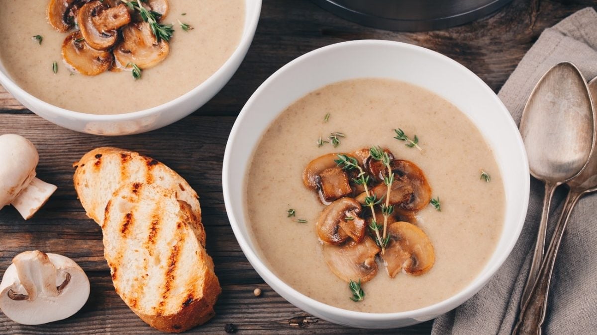 Draufsicht: Zwei helle Schüsseln mit einer pürierten Bohnensuppe mit geschmorten Champignons stehen auf einem Untergrund aus Holz. Auf den Tosch liegen zwei silbererne Suppenlöffel sowie eine geröstete Weißbrotscheibe.