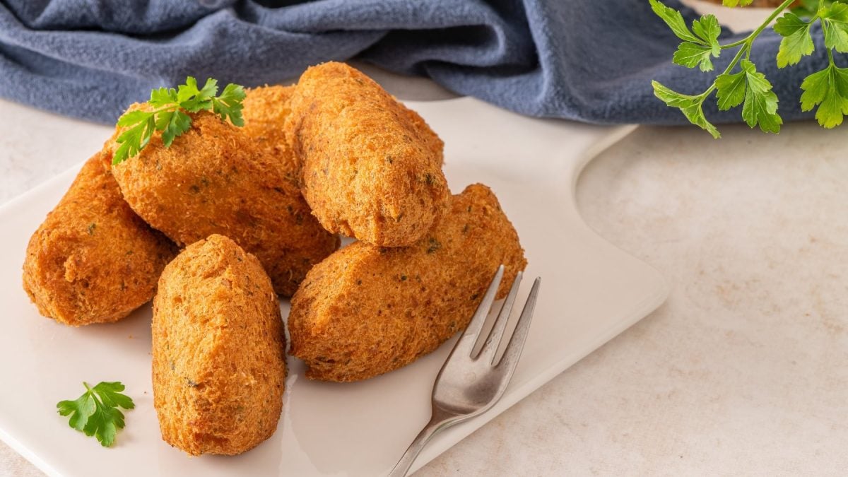 Einige Fischkroketten auf einem weißen Teller mit einem blauen Tuch im Hintergrund. Daneben noch ein Zweig Petersilie.