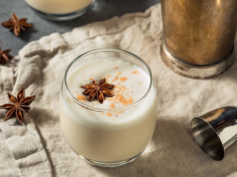 Vanilla Maple Bourbon Milk Punch im Glas mit Sternanis, Barmaß und Shaker auf Tuch und grauem Untergrund. Im Hintergrund ein weiteres Glas. Draufsicht.