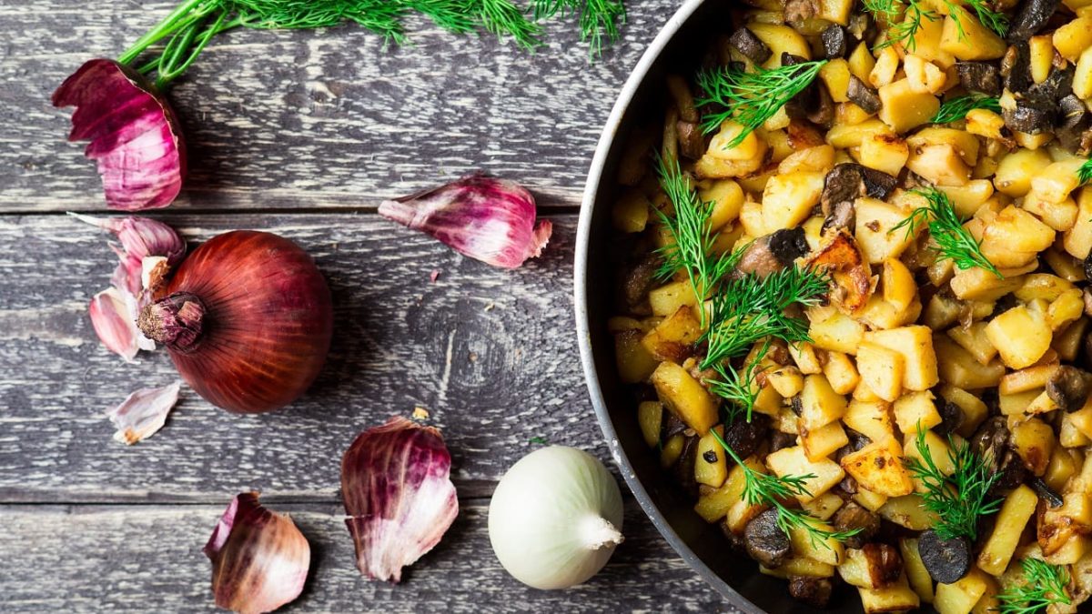 Draufsicht: Auf einer groben Holzplatte liegen eine rote Zwiebel, Zwiebelschalen sowie eine weiße Zwiebel. Daneben steht eine Pfanne mit Bratkartoffeln und Birne.
