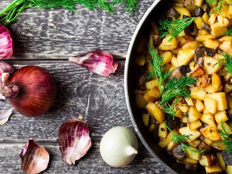 Draufsicht: Auf einer groben Holzplatte liegen eine rote Zwiebel, Zwiebelschalen sowie eine weiße Zwiebel. Daneben steht eine Pfanne mit Bratkartoffeln und Birne.