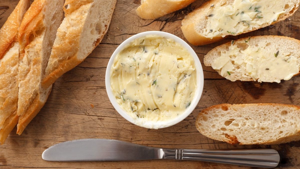 Draufsicht: eine helle kleine runde Schale mit einer kräuterbutter auf einem Untergrund auf Holz. Drum herum lieben aufgeschnittene baguettescheiben.