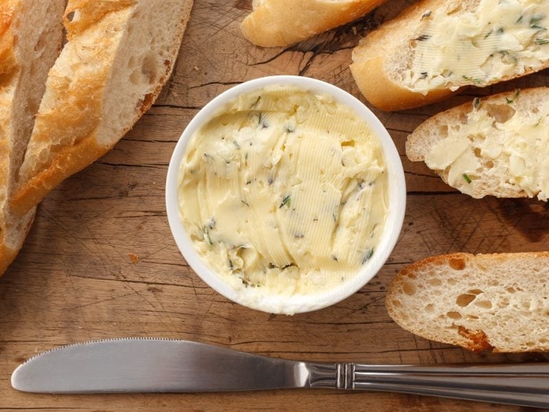 Draufsicht: eine helle kleine runde Schale mit einer kräuterbutter auf einem Untergrund auf Holz. Drum herum lieben aufgeschnittene baguettescheiben.