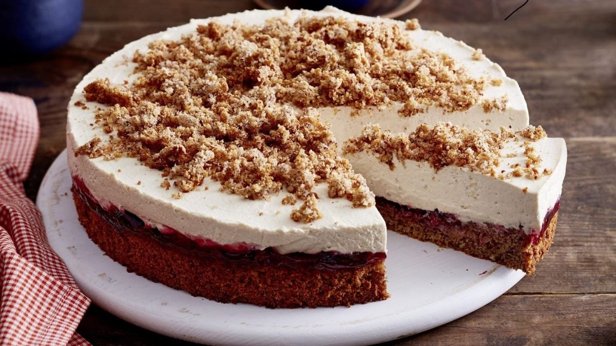 Angeschnittene Torte auf einem weißen flachen Teller auf einem hölzernen Untergrund. Die Torte hat einen braunen Boden und eine helle Cremeschicht, die mit Krümeln dekoriert ist.