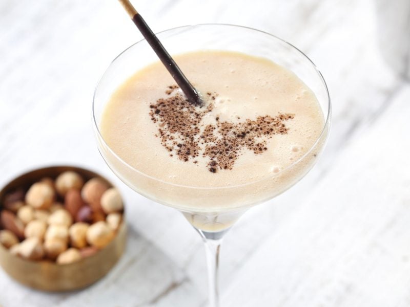 Ein weißer Cocktail im Martiniglas aus dem Buch "Cocktails – die besten Drinks", daneben unscharf eine Schale Nüsse.