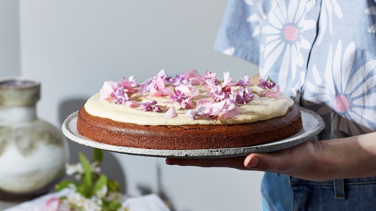 Eine Frau hält eine Kuchenplatte mit einer veganen Dattel-Cashew-Torte, auf der Blüten liegen.
