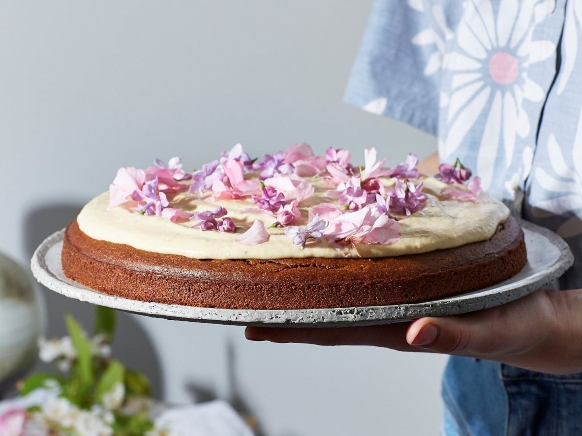 Eine Frau hält eine Kuchenplatte mit einer veganen Dattel-Cashew-Torte, auf der Blüten liegen.