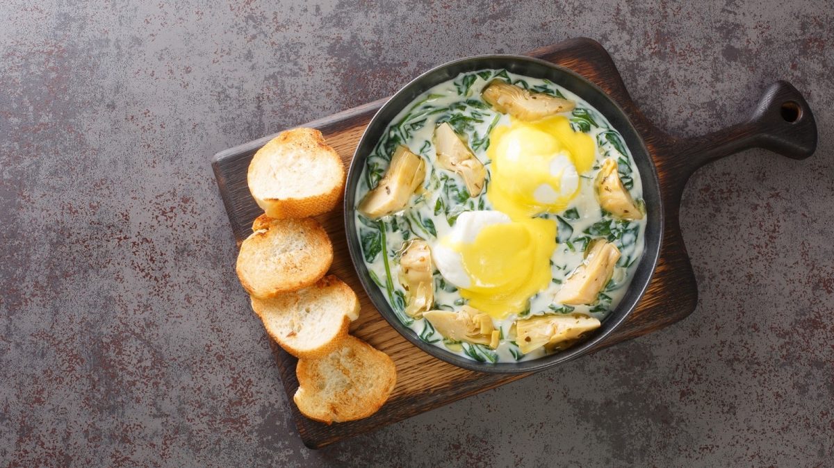 Eine schwarze Pfanne mit Eggs Sardou auf einem Holzbrett.