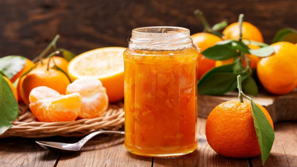 Ein Glas mit Mandarinen-Marmelade vor frischen Mandarinen.