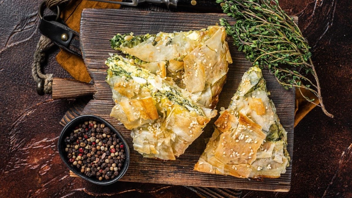 Drei Stücke Spanakopita auf einem Holzbrett neben einer Schale mit Pfefferkörnern.