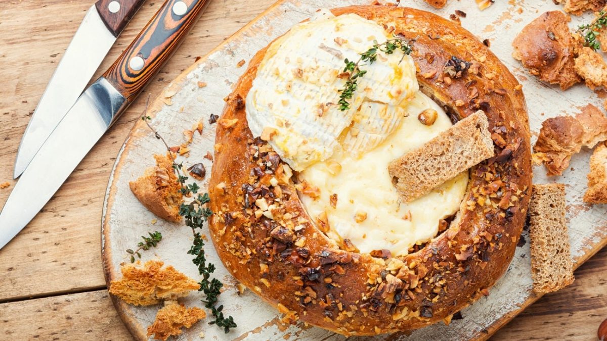 Draufsicht: Ein rundesbrot ist in der Mitte ausgehölt. In dem Loch ist ein zerlassener Camembert-Käse. Auf ihn ist ein Zweig Thymian. Drum herum liegen brotstücke und zwei Messer mit Schäften aus Holz.