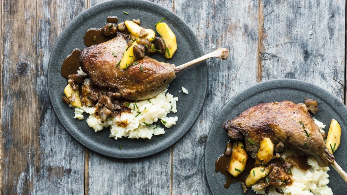Gänsekeulen mit Äpfeln und Maronen auf Reis, serviert auf Keramiktellern auf einem grauen Holztisch.