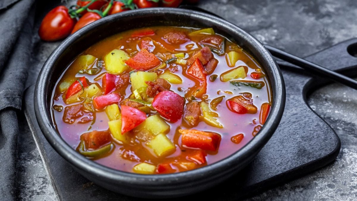 Eine schwarze Schale mitd er Gulaschsuppe auf einem Steinuntergrund und einem schwarzen Schneidebrett. Daneben ein kleiner Strauch mit Kirschtomaten.