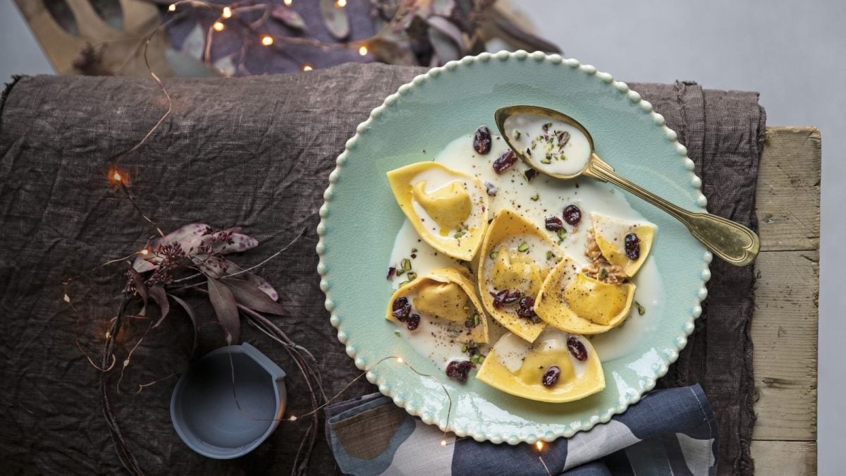Draufsicht: Dunkler, hölzerner Untergrund mit dekorativer Lichterkette und Blumen. Außerdem ein türkisfarbener Teller mit sechs Ravioli in einer cremig hellen Sauce. Garniert mit getrockneten Cranberries und Pistazien.