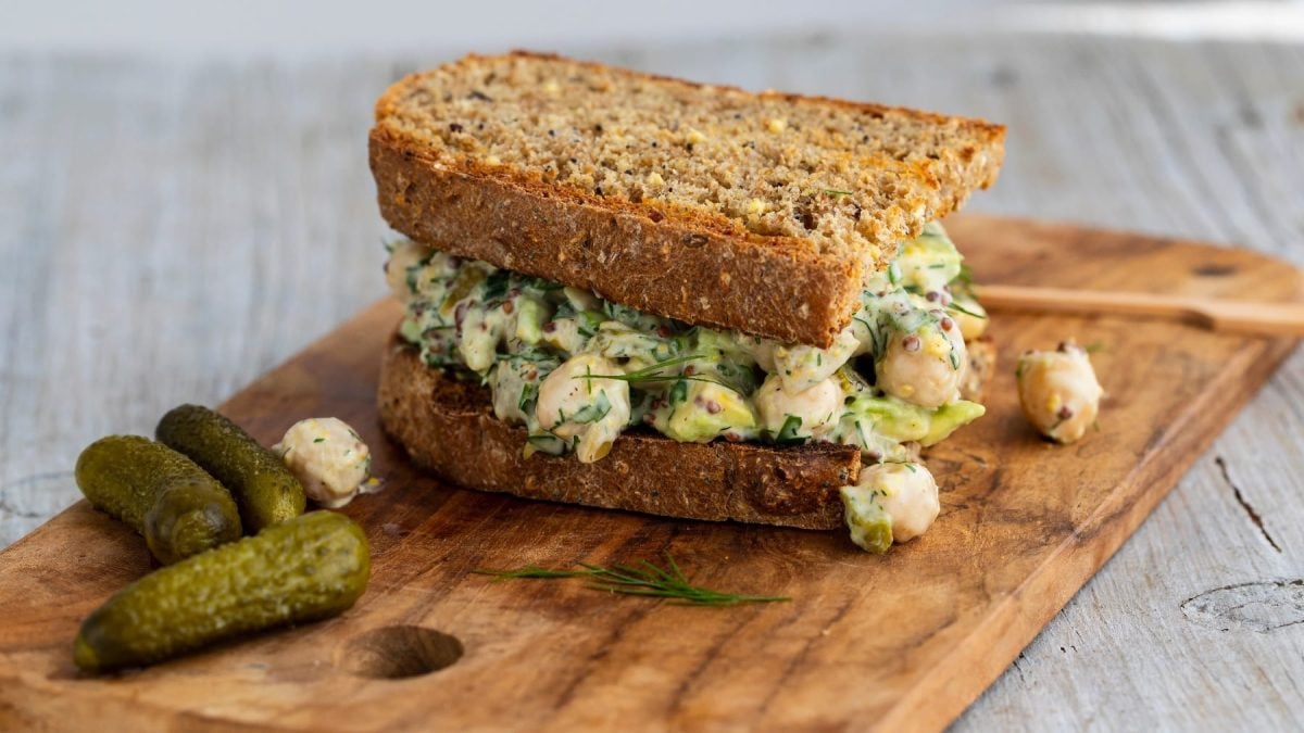 Ein Kichererbsen-Sandwich auf einem Holzbrett mit drei Mini-Gewürzgurken und etwas Dill.