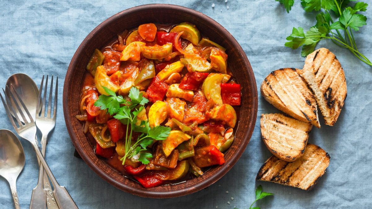 Eine Schüssel Knoblauch-Eintopf mit Röstgemüse in der Draufsicht, daneben getoastetes Brot und Besteck.