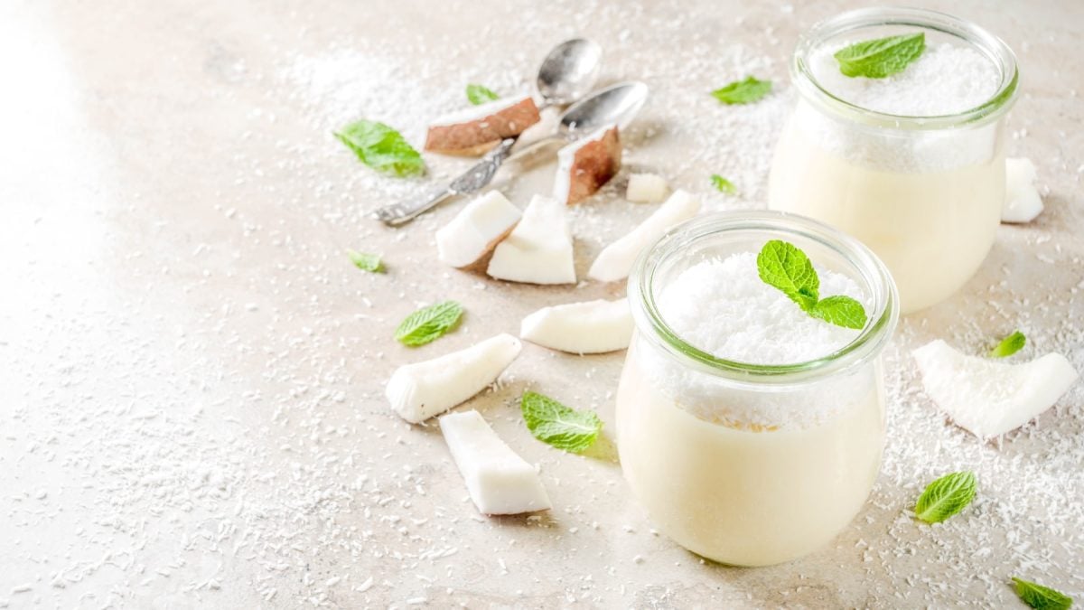 Zwei Gläser mit der weißen Panna Cotta auf grauem Untergrund mit Kokosraspeln und Kokosstückchen.