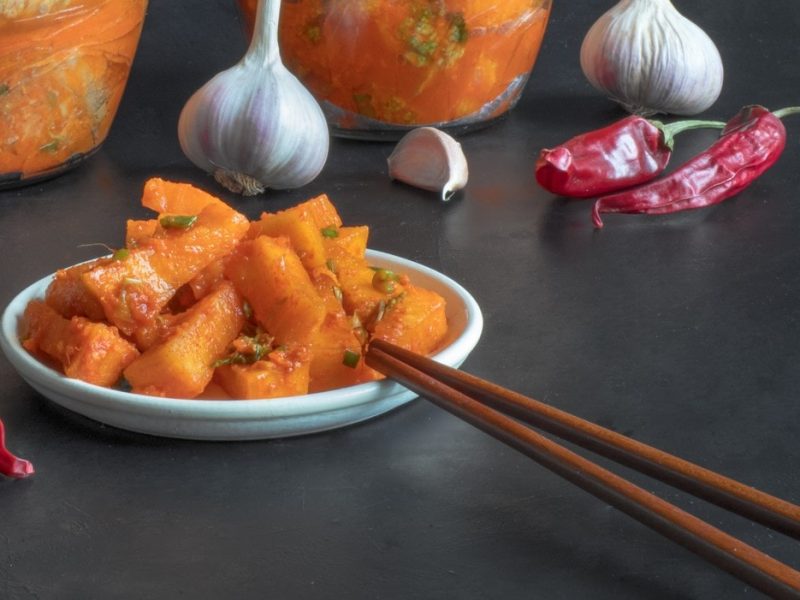 Im Vordergrund das Kürbis-Kimchi auf eine rkleinen weißen Schale. Im Hintergrund zwei Kimchi-Gläser mit Chinakohl.