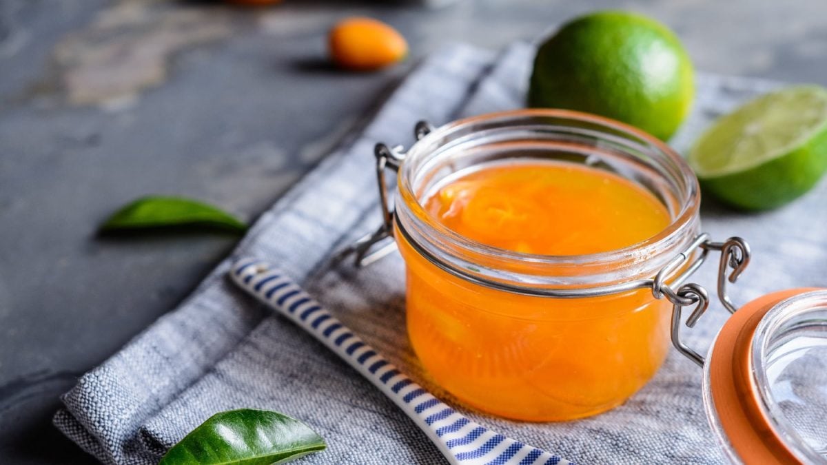 Ein Glas mit der Kumquat-Marmelade auf einem grauen Untergrund. Daneben eine Limette und Kaffirblätter.