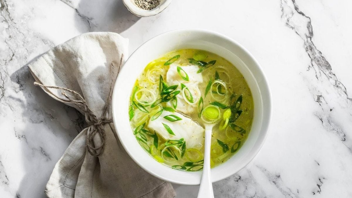 Draugsicht: eine helle Schale mit einer Leichte Lauch-Kabeljau-Suppe steht auf einen Untergrund aus hellem Marmor. In der Suppe ist ein Suppenlöffel.