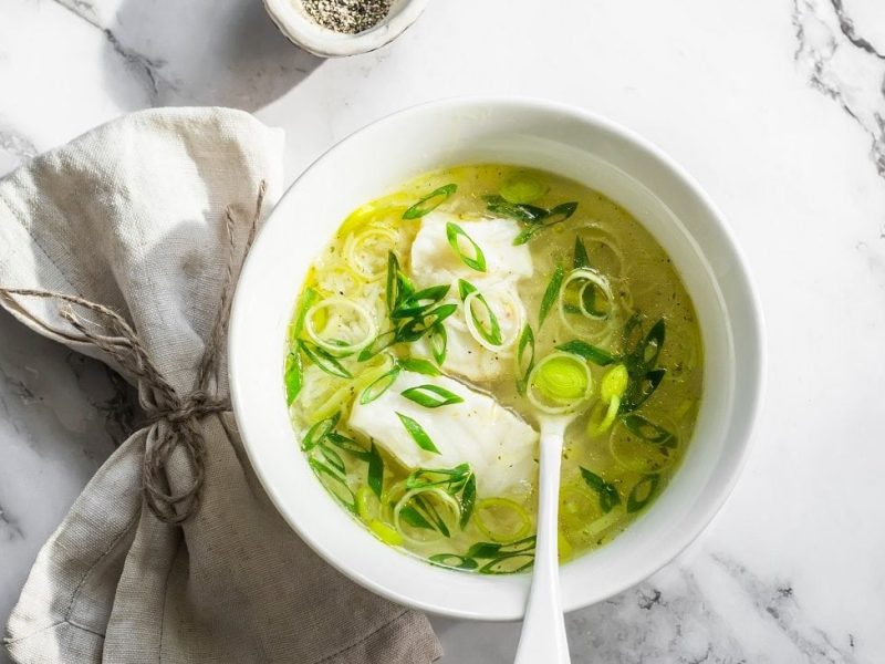 Draugsicht: eine helle Schale mit einer Leichte Lauch-Kabeljau-Suppe steht auf einen Untergrund aus hellem Marmor. In der Suppe ist ein Suppenlöffel.