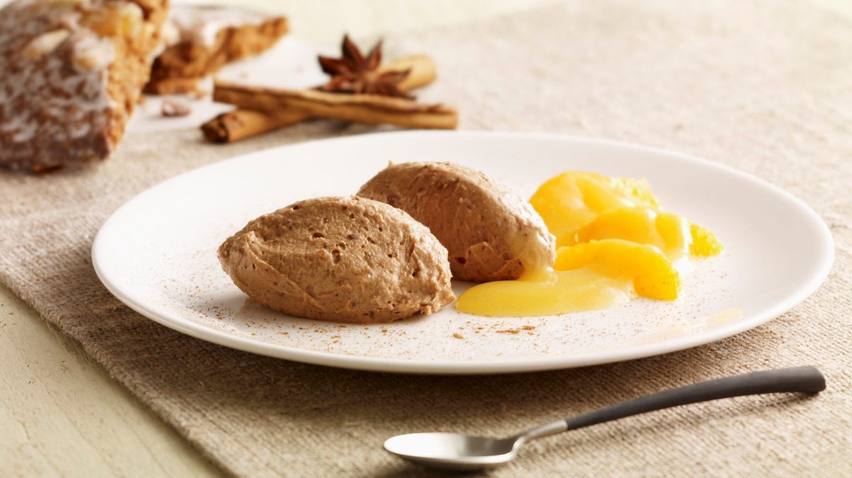 Ein weißer teller auf beigem tisch mit der Lebkuchenmousse und den Orangen. Links daneben Lebkuchen, Zimt und Sternanis