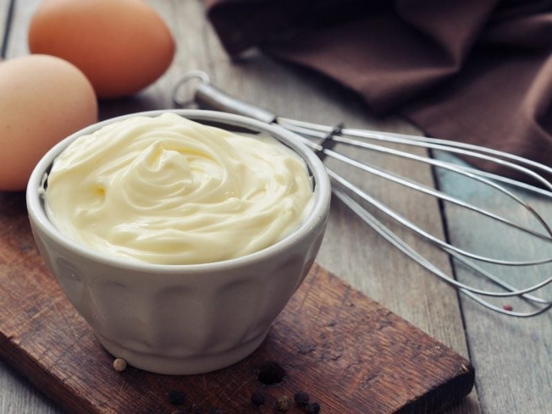 Frontalsicht: Auf einem Holzbrettchen ist eine kleine Schale mit einer dicklichen, hellen Creme. Es handelt sich um eine Mayonnaise. Naneben liegen zwei Eier und ein Schneebesen.