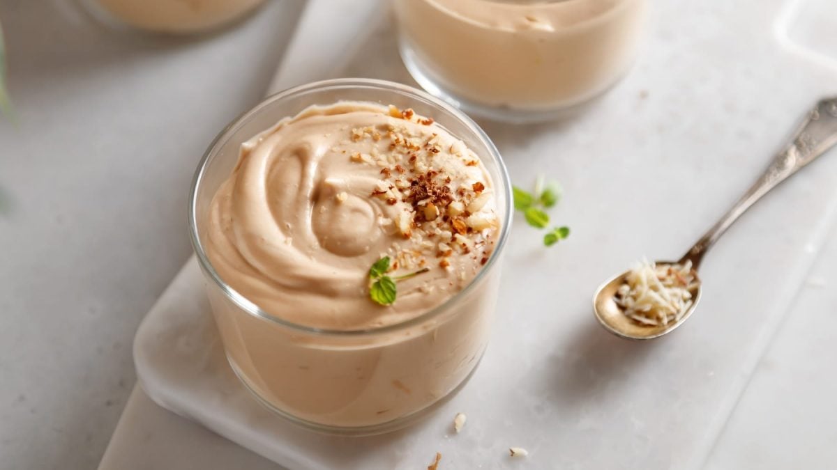 Zwei Gläser mit der hellbraunen Mandel-Mousse auf einem weißen Tisch. mit einem langen, kleinen silbernen Löffel.