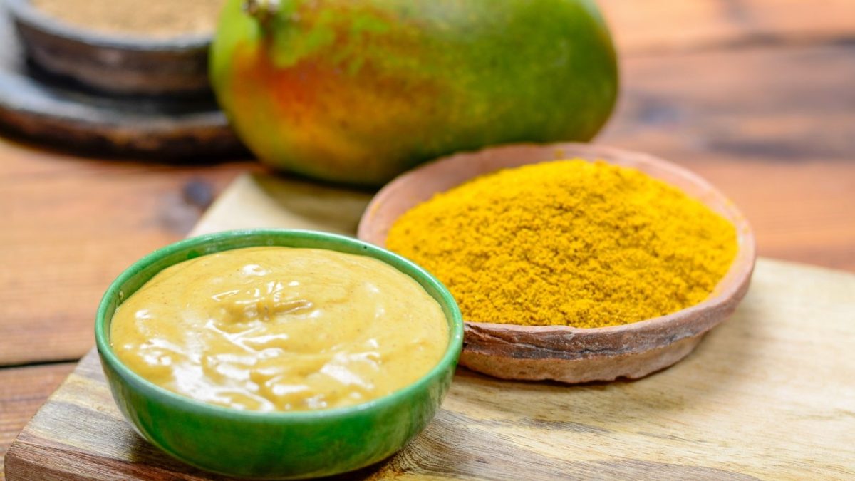 Frontalsicht: drei Schälchen und eine Mango liegen auf einem Untergrund aus Holz. In einem Schälchen ist ein Pulver und in dem nächsten ein senffarbener Dip. Im Hintergrund liegt eine frische Mango.