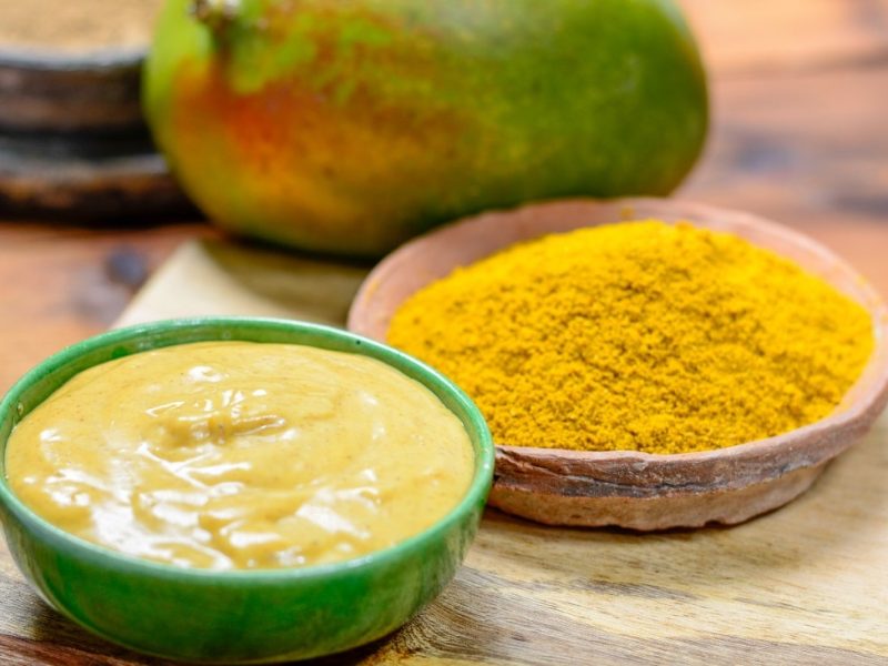 Frontalsicht: drei Schälchen und eine Mango liegen auf einem Untergrund aus Holz. In einem Schälchen ist ein Pulver und in dem nächsten ein senffarbener Dip. Im Hintergrund liegt eine frische Mango.