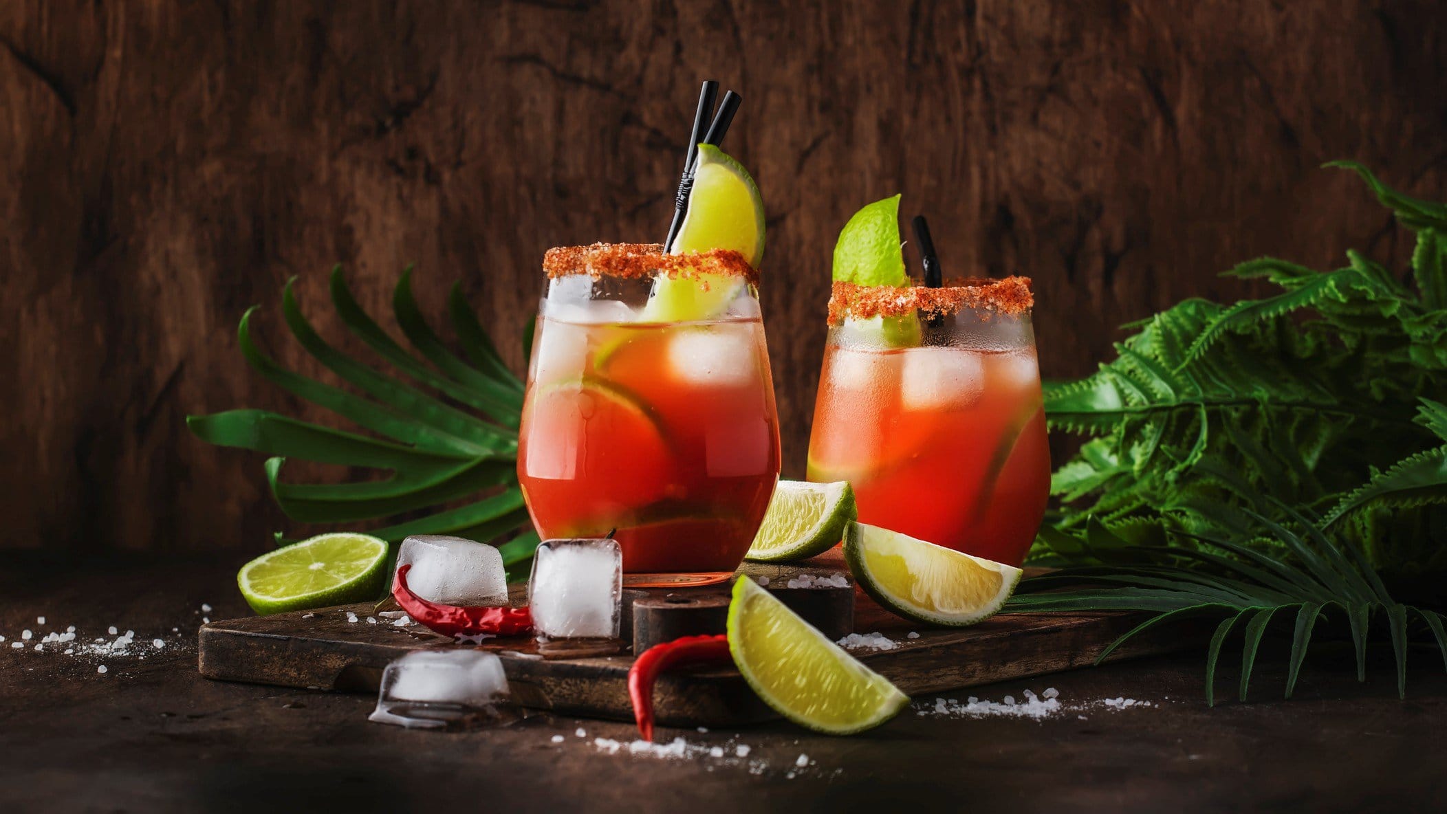 Michelada mexikanischer Biercocktail mit Lager in zwei Gläsern auf dunklem Holz vor dunklem Hintergrund. Im Bild Pflanzen, Limettenspalten, Salz, Chilis und Eiswürfel. Frontalansicht.