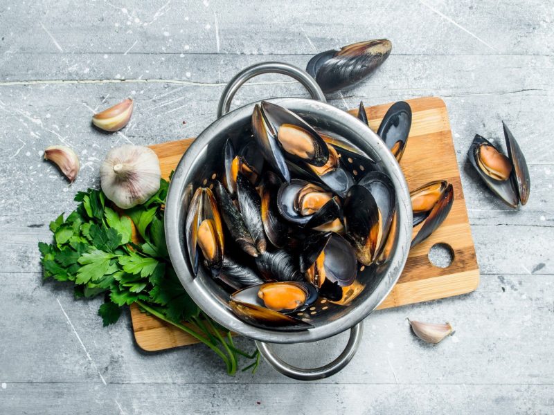 Warum sollte man geschlossene Muscheln nicht essen? Miesmuscheln im Topf auf Holzbrett und grauem Untergrund. Einzelne Muscheln, Knoblauch und Kräuter im Bild. Draufsicht.
