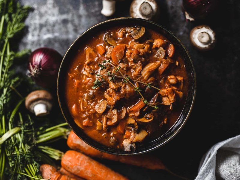 Eine Schüssel No Beef Bourguignon aus dem Kochbuch "Dieses Jahr feiern wir vegan" in der Draufsicht, drumherum frisches Gemüse.