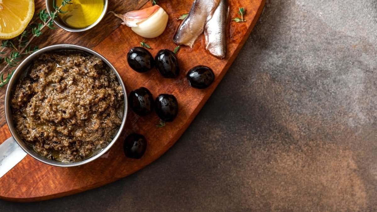 Draufsicht: Auf einem Holzbrettchen, das nur zu einem Teil zu sehen ist, ist ein Schälchen mit einer Oliven-Sardellen-Tapenade. Daneben liegen Sardellenfiltes, schwarze Oliven, ein Stängel Thymian und eine Zitronenscheibe,