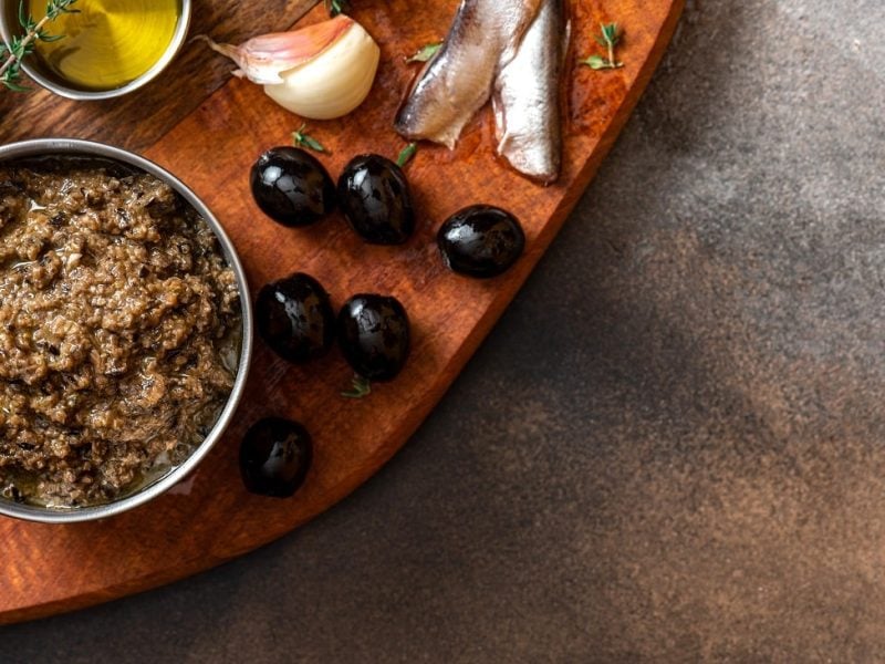 Draufsicht: Auf einem Holzbrettchen, das nur zu einem Teil zu sehen ist, ist ein Schälchen mit einer Oliven-Sardellen-Tapenade. Daneben liegen Sardellenfiltes, schwarze Oliven, ein Stängel Thymian und eine Zitronenscheibe,