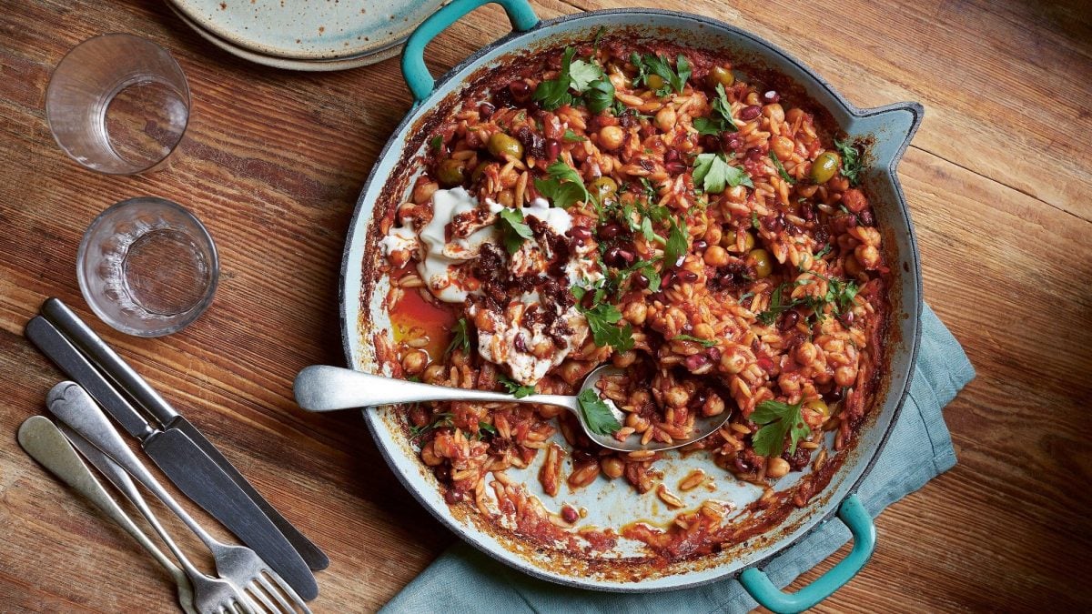 Eine Auflaufform Orzo mit Kichererbsen und Oliven, darüber frische Kräuter, in der Draufsicht.