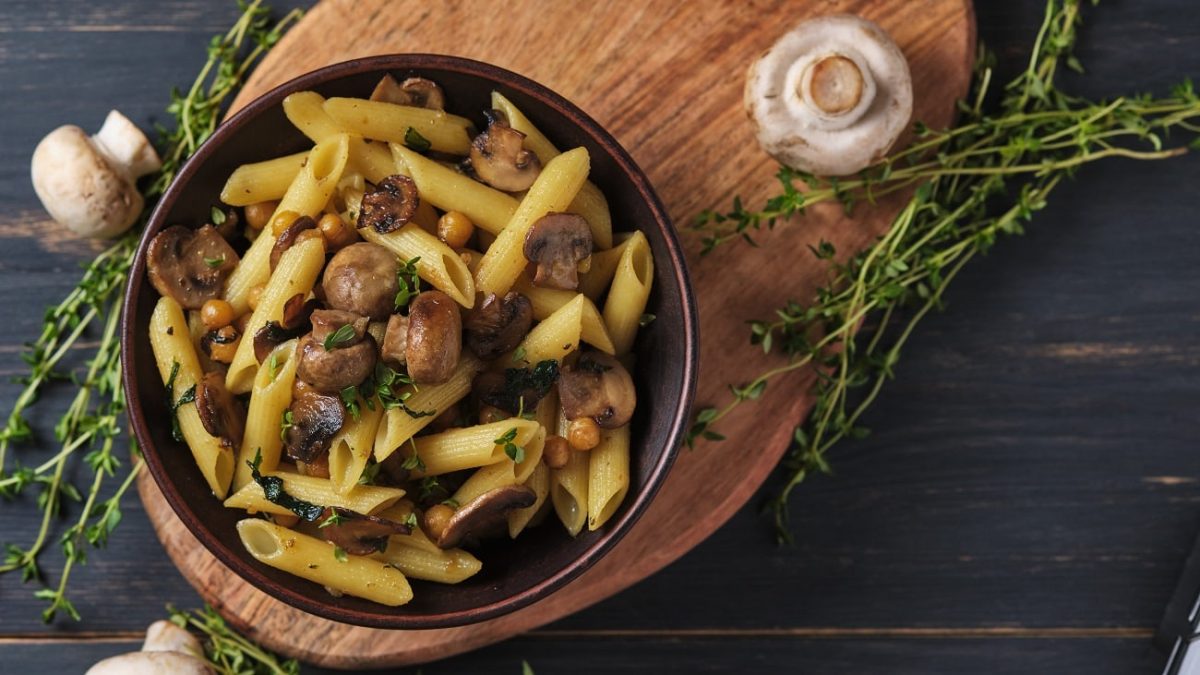 Draufsicht: Auf einem Holzbrettchen steht eine schwarze Schüssel mit Pasta mit Thymian-Champignons. neben dem Brettchen liegen rohe Champignons und mehrere Zweige frischen Thymian.