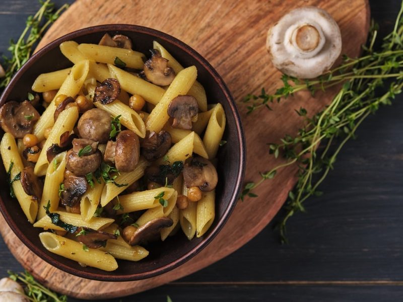 Draufsicht: Auf einem Holzbrettchen steht eine schwarze Schüssel mit Pasta mit Thymian-Champignons. neben dem Brettchen liegen rohe Champignons und mehrere Zweige frischen Thymian.