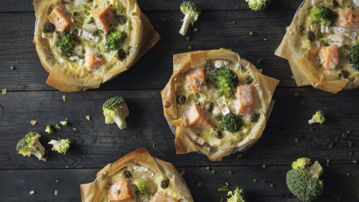 Draufsicht: Dunkler Untergrund mit vier kleinen Quiche. Knuspriger Filoteig gefüllt mit Lachs, Brokkoli und cremigem Käse.