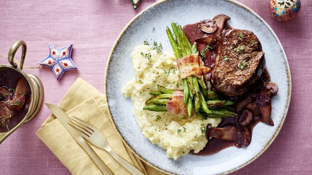 Draufsicht: Rosane Tischdecke mit einem Teller, gelber Servierte und Besteck daneben. Auf dem Teller befindet sich ein Stück Rinderfilet in Rotweinsauce mit Pilzen. Dazu serviert wird Kartoffelpüree und Bohnen im Speckmantel. Alles garniert mit frischen, gehackten Kräutern.