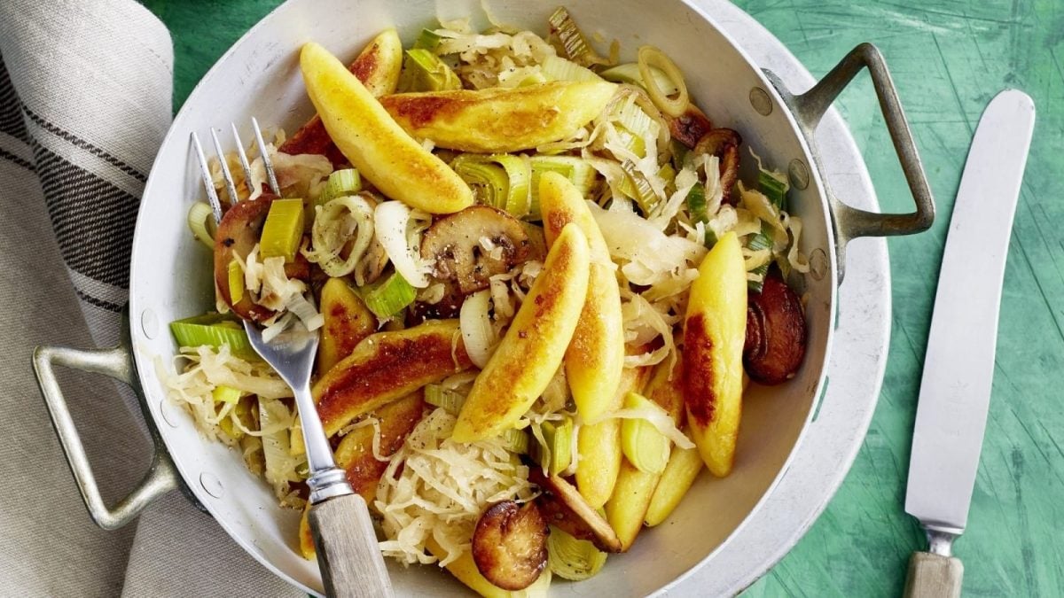 Draufsicht: Grüner hölzerner Untergrund auf dem eine kleine Metallpfanne gefüllt mit gebratenen Schupfnudeln, Sauerkraut, Champignons und frischen Frühlingszwiebeln.