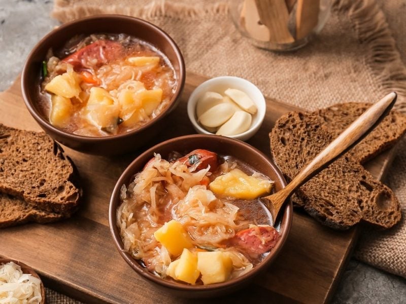 Draufsicht: Zwei dunkle Schalen mit Sauerkrautsuppe und kallser stehen auf einem Holzbrettchen. In einer Suppe ist ein Holzlöffel. Drumherum liegen Brotscheiben, eine helle Schale mit geschältem Knoblauch und ein dunkles Schälchen mit ungeschältem Knoblauchzehen und eine dunkles mit rohem Sauerkraut.