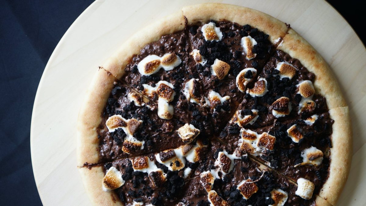 Draufsicht: Dunkler Untergrund mit einem hölzernen Pizzaschieber. Darauf eine Pizza mit Schokoladencreme, Oreo-Stückchen und karamellisierten Marshmallows