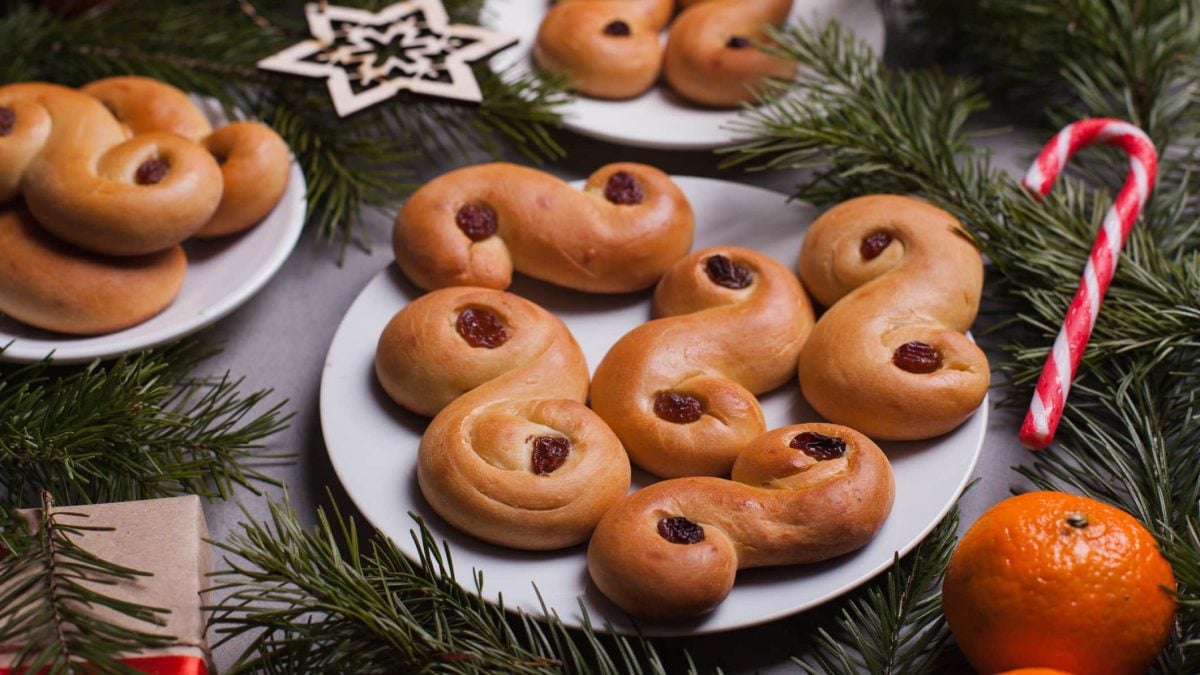 Schwedische Lussekatter auf einem Teller, daneben Tannengrün, Mandarinen und Zuckerstangen.