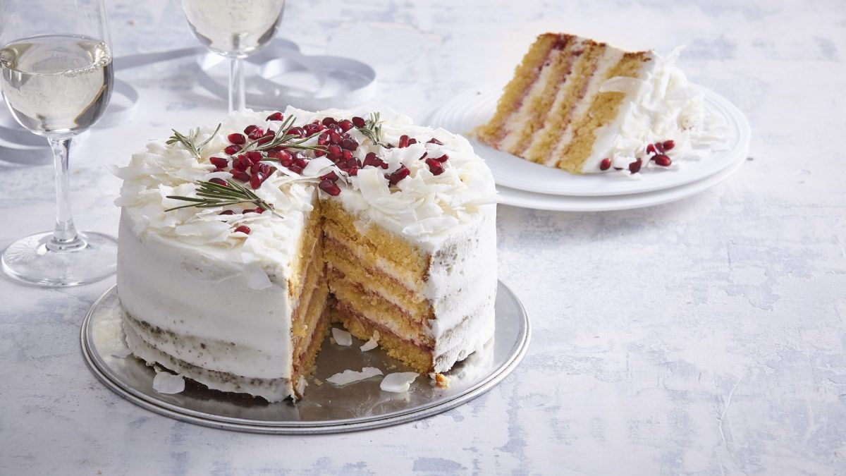Angeschnittener Silvesterkuchen mit Mandeln und Kokos verziert mit Granatapfelkernen und Rosmarin auf einem Teller vor hellem festlichen Hintergrund.