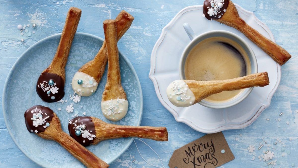 Mehrere Spekulatius-Löffel mit dunkler und weißer Schokolade und Streuseln neben einer Tasse Kaffee auf blauem Teller.