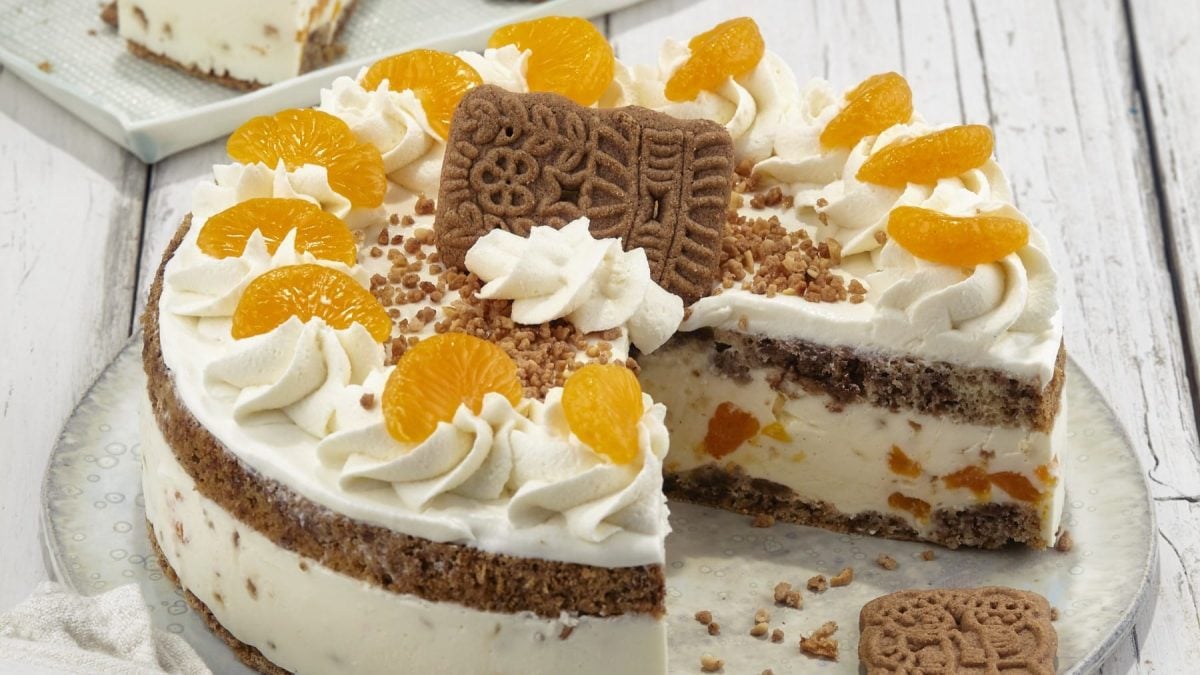 Angeschnittene Spekulatius-Sahnetorte mit Mandarinen auf einem weißen Teller vor hellem Hintergrund.
