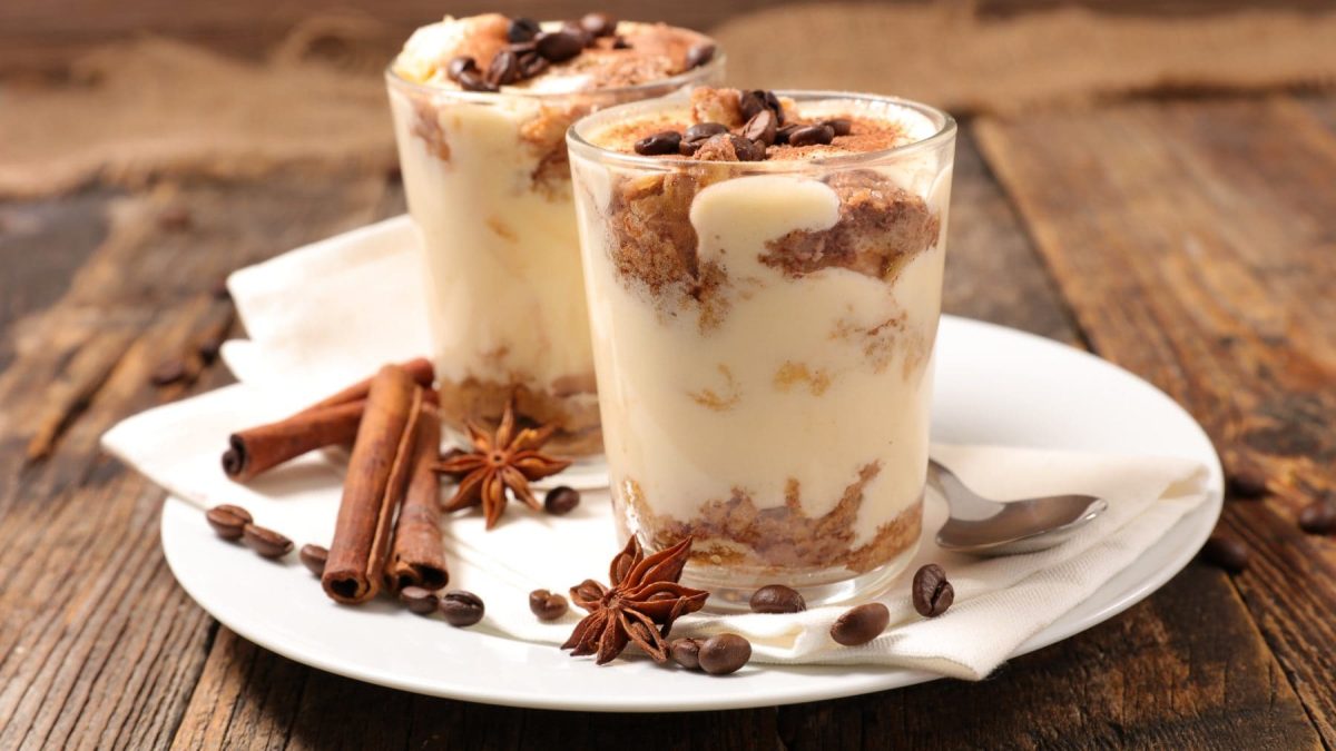 Drei Spekulatius Tiramisu in Gläsern auf Holzbrett aus der Frontale. Auf dem Brett liegen Pinienzapfen, kleine goldenen Kugeln und Nadelzweige.
