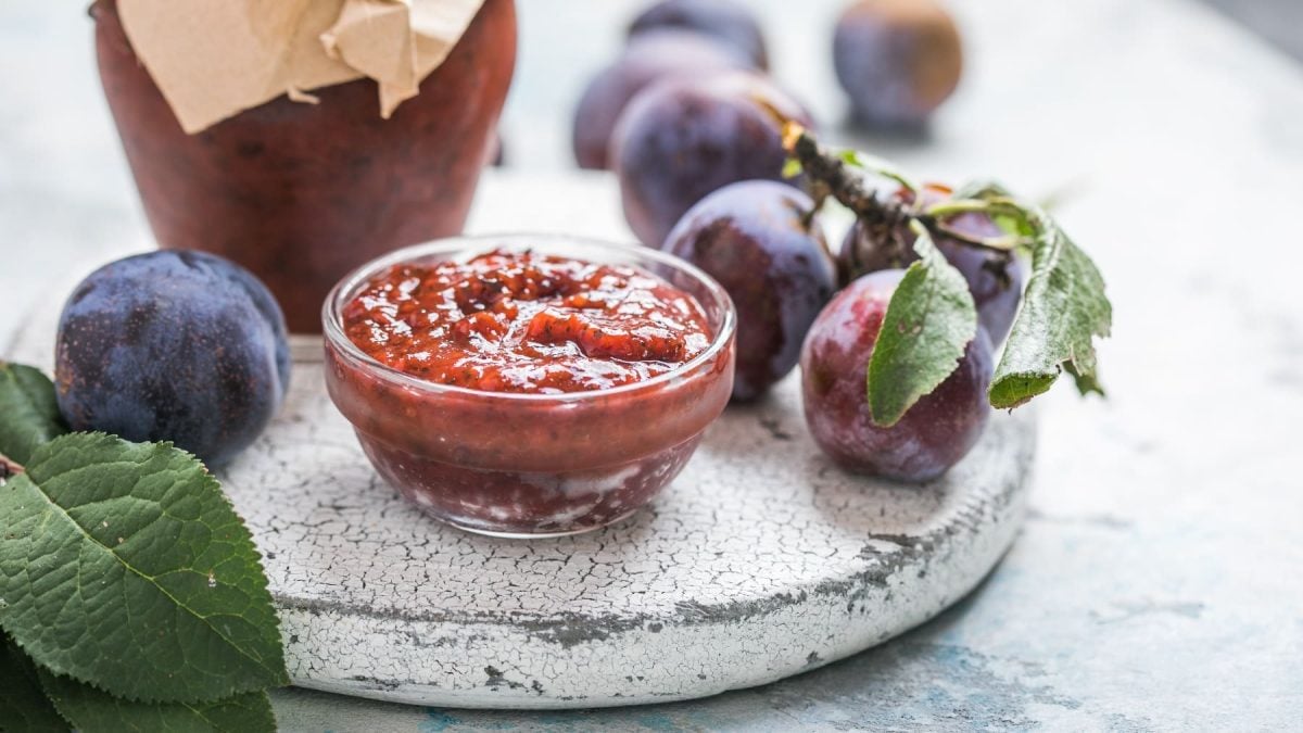 Eine kleine Glasschale mit der Pflauemnsauce auf einer grauen Steinplatte. Im Hintergrund auch frische Pflaumen und ein Glas mit Konfitüre.
